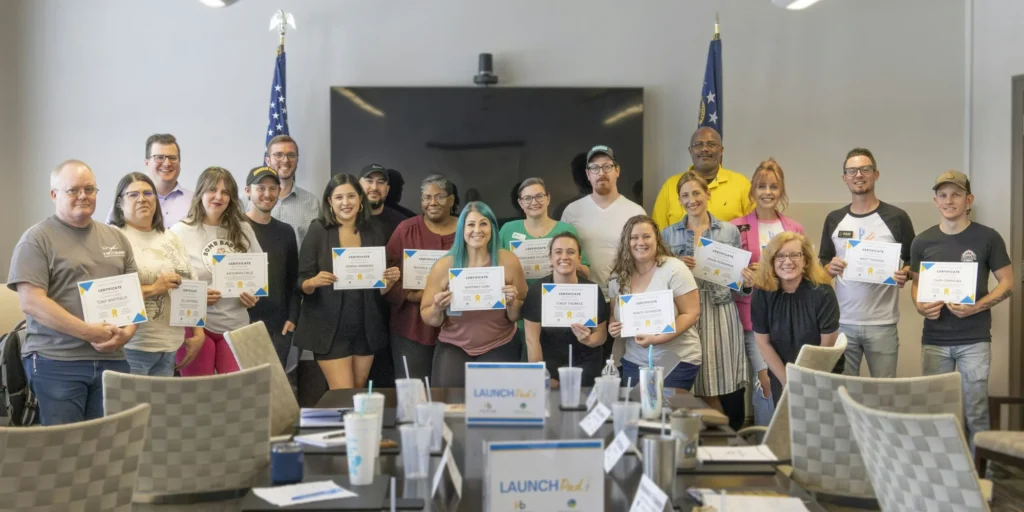 A group of 17 COED launchpad bootcamp graduates smiling, holding certificates in a conference room. They stand in front of a screen and U.S. flags, conveying a celebratory mood.
