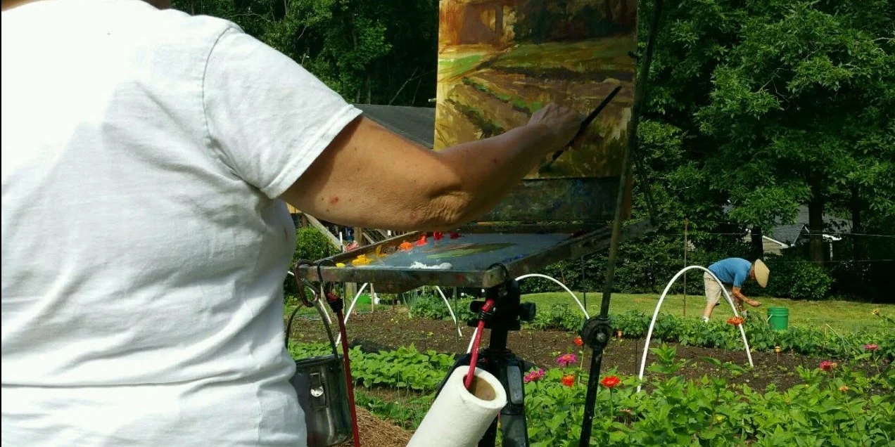A person in a white shirt paints a scenic view of a garden on a canvas using an easel, with vibrant flowers and greenery in the foreground.