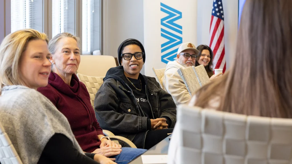 A diverse group of people sits around a conference table, engaged in discussion. They appear friendly and attentive, with an American flag in the background.