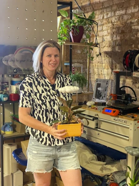 A smiling person in a zebra-patterned shirt holds a potted plant in a vibrant workspace with tools and colorful decor, conveying creativity and warmth.