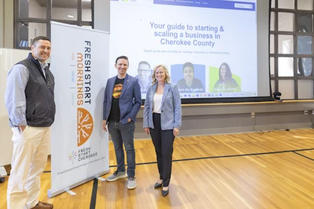 Three people smile in front of a large screen displaying a business guide for Cherokee County. A sign reads "Fresh Start Mornings." The setting is bright and professional.