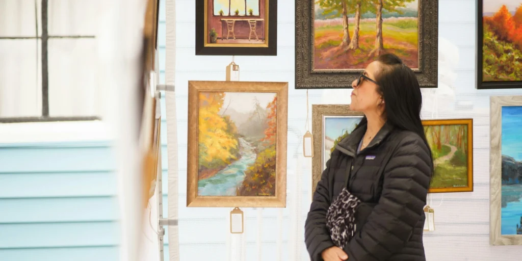 A woman in a black jacket views colorful landscape paintings at an art gallery. The scene conveys a calm, contemplative atmosphere.
