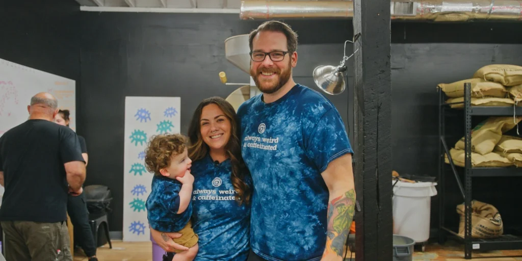 Smiling family in tie-dye shirts that say, "always weird & caffeinated." They stand in a cozy room with shelves, conveying joy and togetherness.