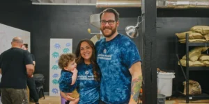 Smiling family in tie-dye shirts that say, "always weird & caffeinated." They stand in a cozy room with shelves, conveying joy and togetherness.