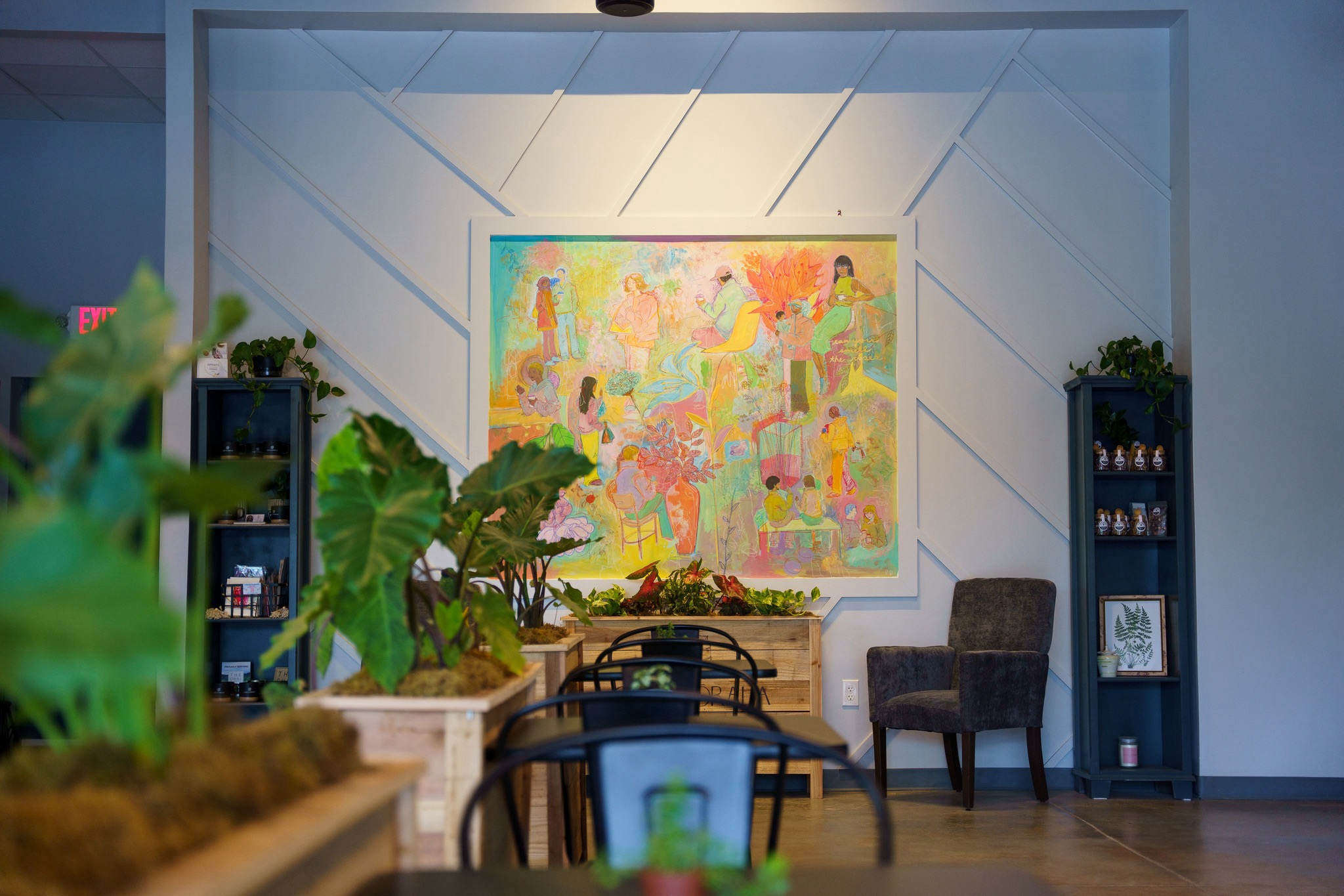 Interior of a cozy, plant-filled coffee shop with colorful wall art, black chairs, wooden planters, and modern decor.