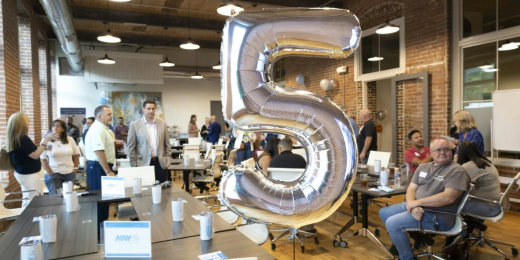 A large silver balloon shaped like the number 5 is in the foreground of a bright office space with exposed brick walls. People are mingling and sitting at tables, creating a lively and celebratory atmosphere.
