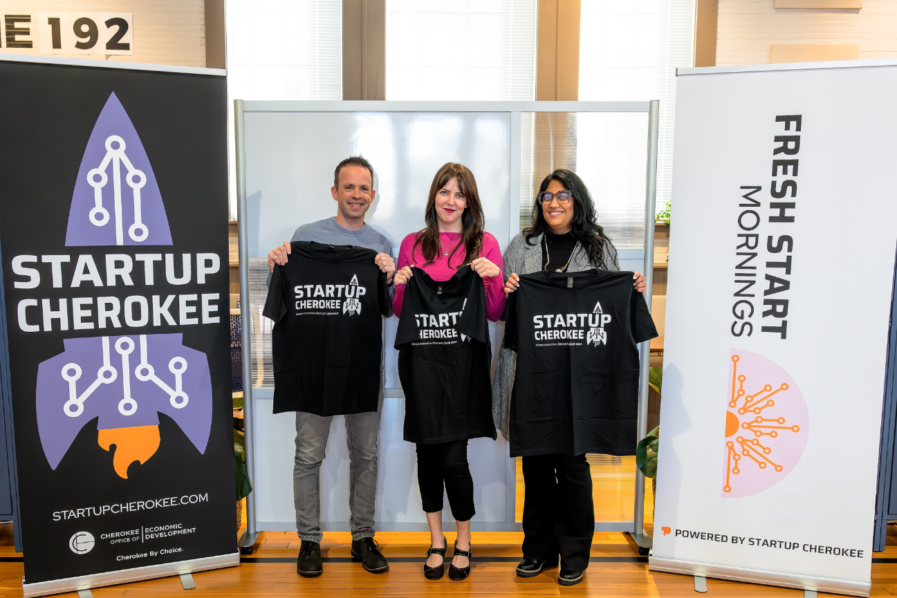 Three people holding Startup Cherokee T-shirts, standing between banners for Startup Cherokee and Fresh Start Mornings at an entrepreneur networking event.