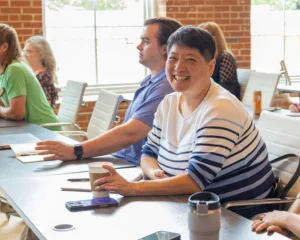 A cheerful individual in a striped sweater smiles while holding a coffee cup. They're seated at a conference table with others, in a well-lit room.